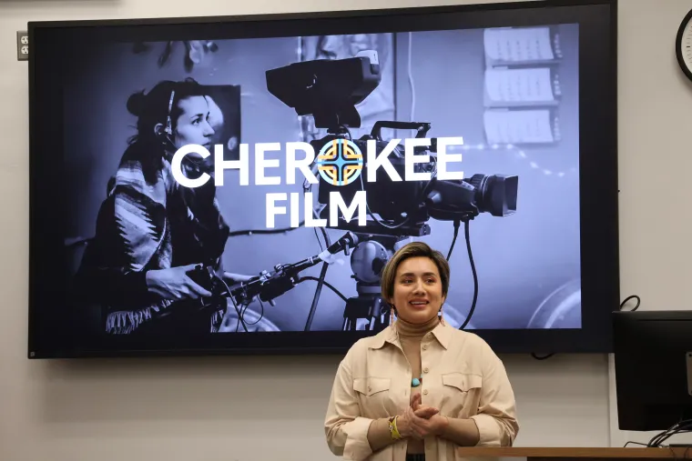 Maggie Cunningham wears a cream-colored shirt open over a gold turtleneck and large turquoise necklace. A large screen behind her features a black and white image of a young woman working a ciematic camera with the text "Cherokee Film" overlaid in white.